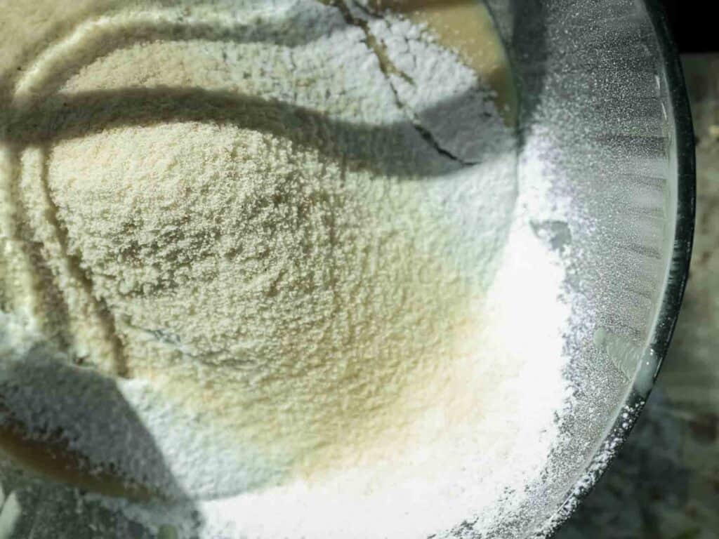 A close-up view of white powdery flour in a clear glass mixing bowl. Natural light casts soft shadows across the textured surface, highlighting its smooth, granular appearance.